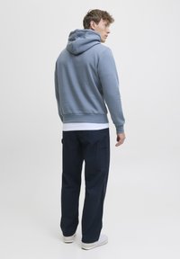 Uomo in piedi, che indossa un hoodie blu, una maglietta bianca, pantaloni cargo blu scuro e sneakers bianche, con uno sfondo chiaro e uniforme.