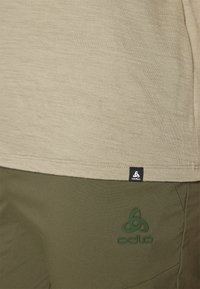 Camisa de manga corta beige con un acabado texturado, que presenta una etiqueta de marca negra, combinada con pantalones verde oliva que tienen un detalle de logo en relieve.