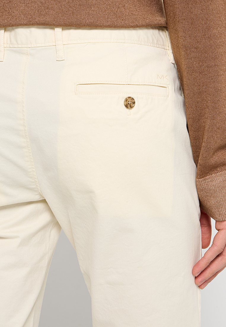 Pantalons blancs avec une poche arrière ornée d'un bouton rond et décoré ; tissu texturé et coupe ajustée visible ; associés à un pull marron.