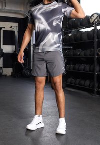 Conjunto deportivo que incluye una camiseta gráfica gris y negra y pantalones cortos grises. El calzado son zapatillas blancas con detalles negros. Entorno de gimnasio con pesas.