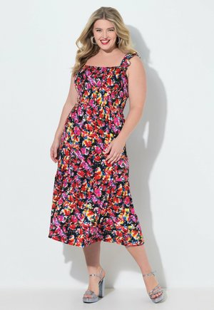 Smilende kvinde med langt blondt hår iført en farverig blomsterprintet midi-kjole og sølvfarvede platformhæle mod en ensartet lys baggrund.