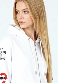 Sweat à capuche blanc zippé avec un intérieur de capuche doublé, présentant un texte noir sur une manche et un logo rouge. Tissu lisse avec des accents en métal argenté.