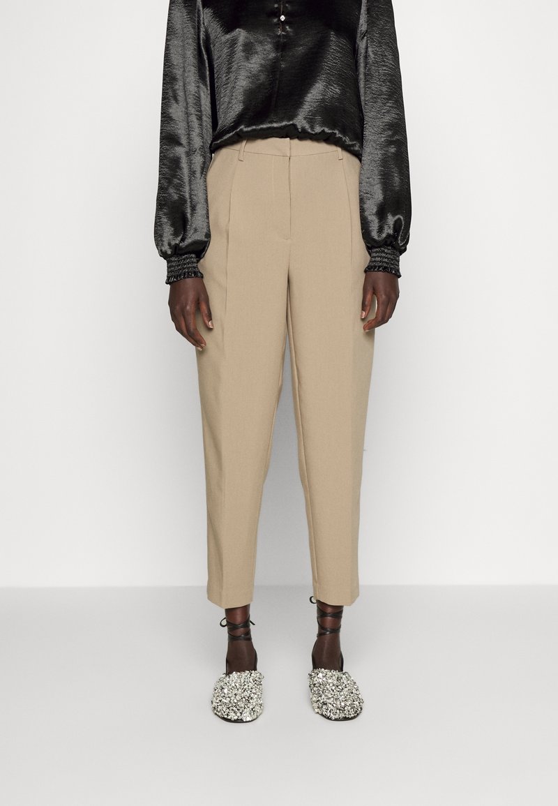 Femme portant un pantalon beige sur mesure longueur cheville, un chemisier noir brillant à manches longues et des chaussures plates ornées avec des brides à la cheville.