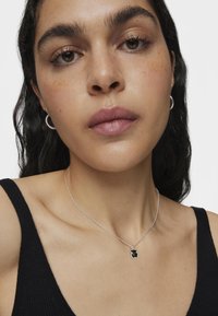 Collar de plata con un colgante negro en forma abstracta, llevado sobre piel desnuda y contrastado con una camiseta sin mangas negra.