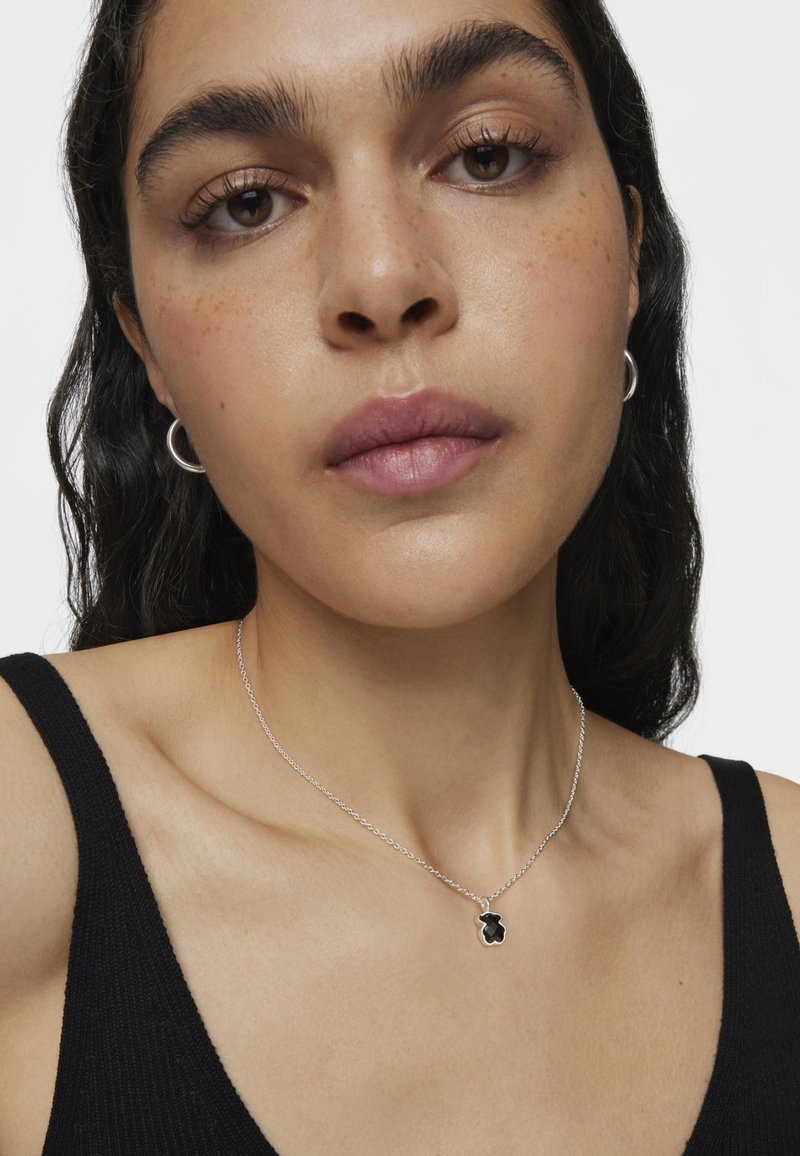 Collar de plata con un colgante negro en forma abstracta, llevado sobre piel desnuda y contrastado con una camiseta sin mangas negra.
