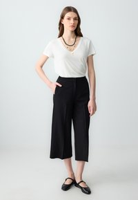 T-shirt blanc à col en V associé à un pantalon noir à jambes larges. Le modèle porte des ballerines noires et un collier à plusieurs couches. Textures lisses et design minimaliste.