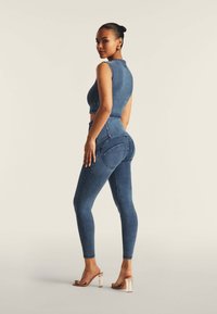Denim-asuste, joka koostuu hihattomasta lyhyestä paidasta ja korkeavyötäröisistä skinny-farkuista, keskianssin sinisessä pesussa, näkyvillä ompeleilla ja sivutaskuilla.
