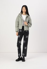 Doudoune vert clair avec capuche, fermeture éclair sur le devant et poignets côtelés ; associée à un pantalon en denim noir à motifs d'étoiles et des chaussures noires.