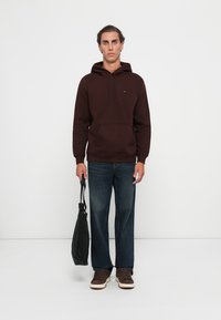 Brown hoodie with kangaroo pocket, drawstring hood, paired with blue jeans and brown sneakers. Model holds a black tote bag.