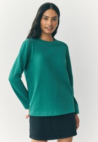 Junge Frau mit langen dunklen Haaren, die ein teal-farbenes Langarmshirt und einen schwarzen Jeansrock trägt, vor einem einfachen hellen Hintergrund.
