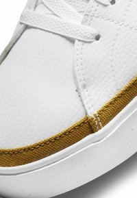 Sneaker bianche in pelle texture con un accento in tessuto dorato a metà suola, dotate di lacci piatti bianchi e dettagli cuciti.