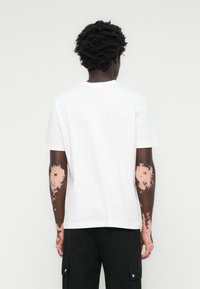Weißes Baumwoll-T-Shirt mit Rundhalsausschnitt, kurzen Ärmeln und geradem Saum, getragen mit schwarzen Cargo-Hosen. Die Rückansicht hebt das Design und die Passform hervor.