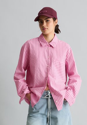 Junge Person trägt eine kastanienbraune Kappe und ein übergroßes rosa gestreiftes Hemd mit Knöpfen, die Hände in den Taschen einer hellblauen Jeans.