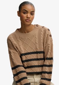 Beige kabelgebreide sweater met zwarte horizontale strepen, voorzien van een ronde halslijn en decoratieve knopen op de linkerschouder.