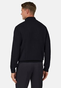 Czarny sweter dzianinowy o wypukłym wzorze, z kołnierzykiem, ściągaczami przy mankietach i u dołu, zaprezentowany z tyłu, w połączeniu z ciemnymi spodniami.