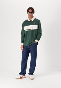 Mladý muž nosí tmavé sluneční brýle, zeleno-bílý dlouhý rukáv polo tričko s textem "Prosím přemýšlejte", modré džíny a bílé tenisky s červenými pruhy.