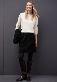 Suéter de punto crema, falda de pana negra, medias oscuras y botas burdeos hasta la rodilla. Texturas suaves y características de diseño simples.