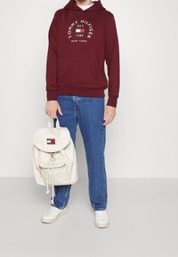 Felpa maroon con logo bianco, jeans azzurri chiari e sneakers bianche. Porta uno zaino crema con branding e una tasca con zip.