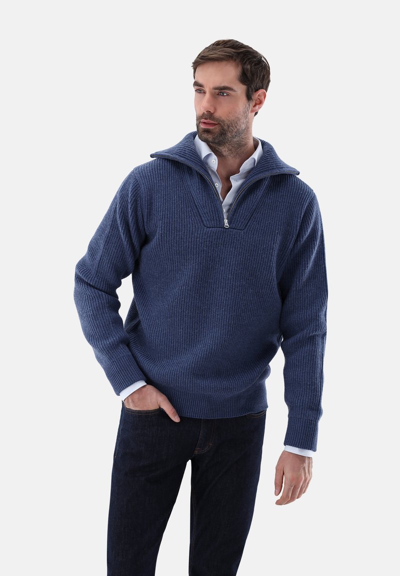 Marineblaues geripptes Sweatshirt mit tiefem Reißverschlusskragen, langen Ärmeln und taillierter Passform; über einem weißen Hemd mit Kragen und dunklen Jeans getragen.