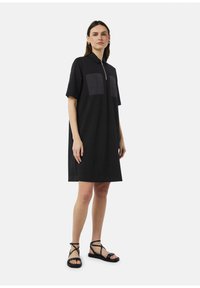 Zwarte polo jurk met een rits aan de voorkant en twee rechthoekige zakken. Korte mouwen en een ontspannen pasvorm. Gecombineerd met zwarte sandalen met bandjes.