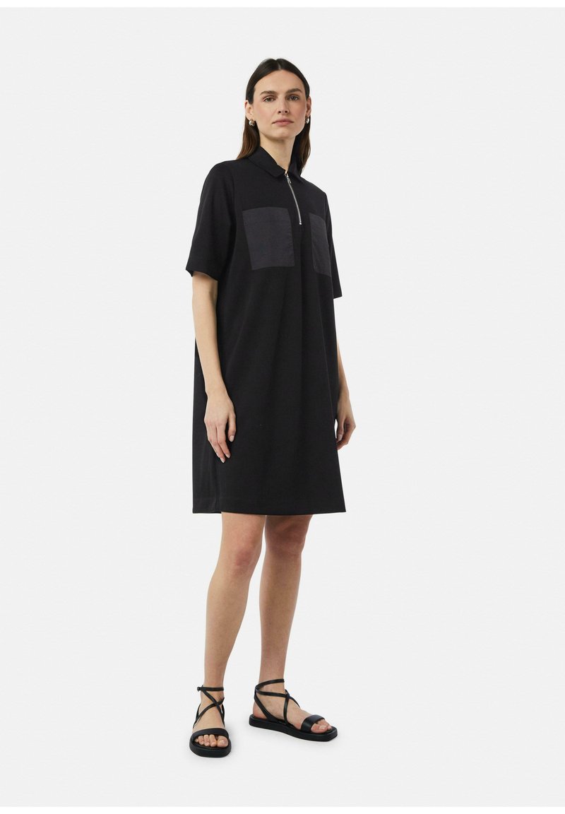Zwarte polo jurk met een rits aan de voorkant en twee rechthoekige zakken. Korte mouwen en een ontspannen pasvorm. Gecombineerd met zwarte sandalen met bandjes.