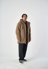 Braune, kapuzenähnliche Außenjacke mit einem Frontreißverschluss, mehreren Taschen und einer lockeren Passform, kombiniert mit schwarzen Hosen und Sneakers.