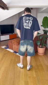 T-shirt azul-marinho com impressão gráfica nas costas, combinada com calções de ganga desbotados. Usado com ténis prateados e meias brancas. Ambiente interno.