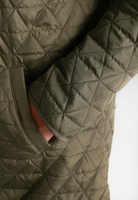Quilted olive-green jacket with diamond pattern, featuring a textured fabric and side pocket, showcasing a fitted silhouette and clean lines.