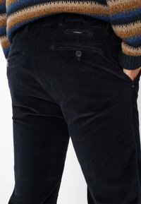 Donkerblauwe corduroy broek met verticale ribbels, voorzien van twee achterzakken, waarvan één met een knoopsluiting, en zijzakken voor de handen.