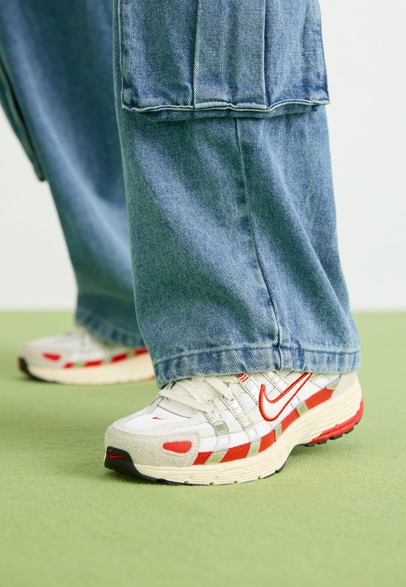 Weiße und rote Nike-Sneaker mit grauen Akzenten, ausgestattet mit einem strukturierten Obermaterial, Schnürsenkeln und einer gepolsterten Sohle, kombiniert mit lockeren blauen Jeanshosen.