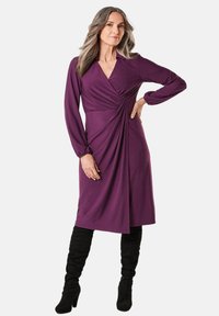 Lila Wickelkleid mit langen Ärmeln, das Raffungen in der Taille und einen Kragen aufweist. Kombiniert mit schwarzen Overknee-Stiefeln. Glatte Textur.