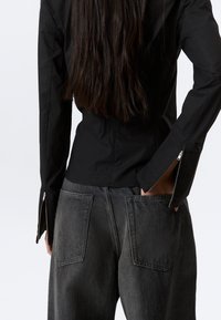 Personne aux longs cheveux foncés portant une veste noire ajustée avec des manches zippées et un jean gris ample avec de grandes poches arrière, main dans la poche.