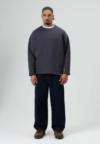 Grijs oversized sweatshirt met een ronde hals, gedragen over een wit shirt, gecombineerd met donkere denim broek en bruine veterschoenen.