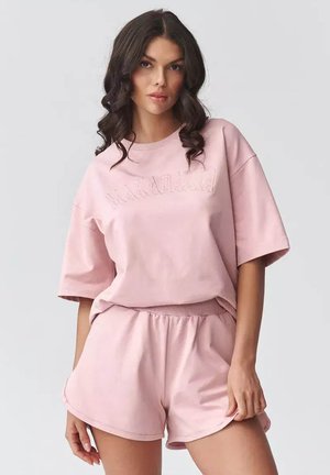 Kobieta o ciemnych, kręconych włosach, ubrana w jasnoróżowy, duży top z krótkim rękawem i dopasowane, wysokie szorty, stojąca na jednolitym tle.