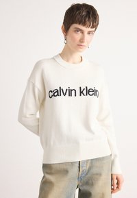 Hvit strikkegenser med svart tekst "calvin klein" på forsiden, med ribbestrikket halslinning og mansjetter, kombinert med lyseblå jeans.