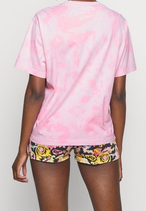 Persoon die een roze tie-dye T-shirt en zwarte korte broek met bloemenpatroon draagt, staand met de rug naar de camera tegen een effen achtergrond.