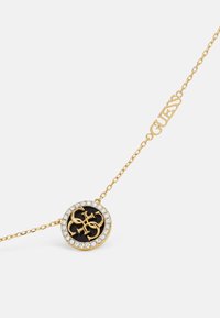 Collana d'oro con pendente circolare, caratterizzato da un centro nero, circondato da strass trasparenti, e un'etichetta 'GUESS' attaccata alla catena.