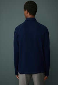 Navy pullover with a textured knit, raglan sleeves, and a well-defined collar. The hem is slightly curved, paired with light gray trousers.