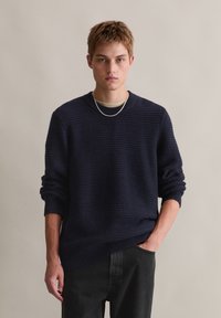 Marineblauer strukturierter Pullover mit Rundhals und gerippten Bündchen, kombiniert mit dunklen Hosen. Gefeaturet ist eine silberne Kettenhalskette.