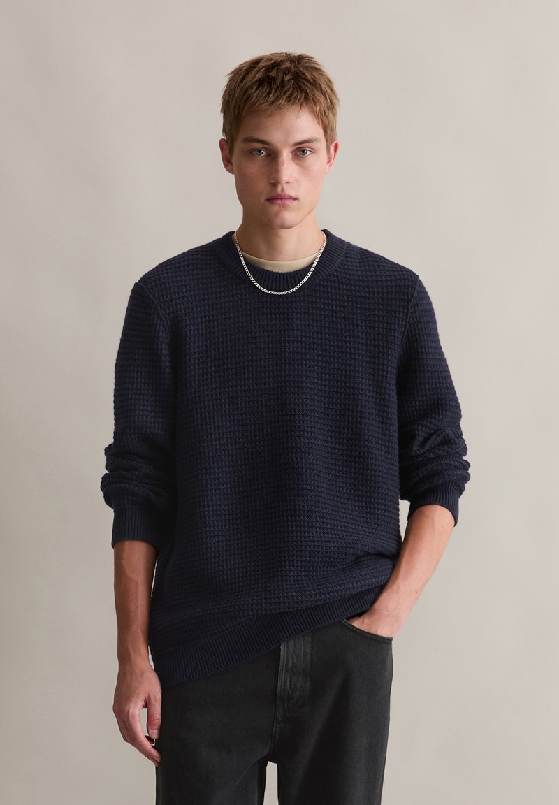 Marineblauer strukturierter Pullover mit Rundhals und gerippten Bündchen, kombiniert mit dunklen Hosen. Gefeaturet ist eine silberne Kettenhalskette.