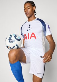 Hvid Nike fodboldtrøje med grå detaljer, prydet med et fremtrædende rødt "AIA" logo. Blå knæhøje sokker. Spiller holder en mønstrede fodbold.