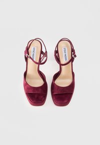 Burgundi zomšos aukštakulniai sandalai su kvadratiniu pirštu, kulkšnies dirželiu ir lygiu smėlio spalvos vidpadžiu su prekės ženklu "Steve Madden."