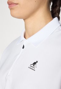 Balta polo marškinėliai su plokščiu apykaklė, ant kairiojo krūtinės ploto pavaizduotas juodas kengūros logotipas ir tekstas „KANGOL J.LINDEBERG“. 