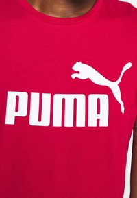 Personne portant un t-shirt rouge avec un logo Puma blanc représentant un puma sautant et un grand texte du nom de la marque sur la poitrine.
