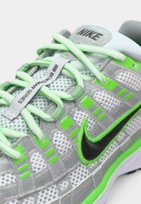 Sive atletske cipele s zelenim naglascima, mrežasti gornji deo, dizajn sa vezicama, crni Nike znak i teksturirani detalji za prianjanje i podršku.
