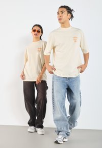 Two models wear oversized, cream t-shirts with orange graphics. One wears dark baggy jeans, the other light blue jeans and sneakers.