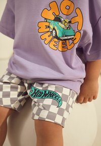 Lila T-Shirt mit einem bunten "Hot Wheels"-Grafikdruck und karierten grauen und weißen Shorts mit einem türkisen "Hot Wheels"-Logo.