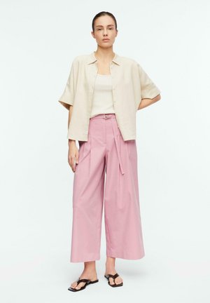 Frau steht, trägt ein beiges Kurzarm-Hemd mit Knöpfen, cremefarbenes Tanktop, hochgeschnittene rosa weite Hose und schwarze Sandalen auf weißem Hintergrund.