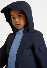 Navy and light blue children's jacket with a hood, featuring a front zipper, patch pocket, and textured fabric. Accented with logo detail.
