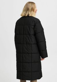 Melnā polsterētā mēteli ar garu siluetu, ar quilted dizainu, augstu apkakli un platām piedurknēm. Gluda, matēta virsma, bez redzamām kalpošanas daļām.